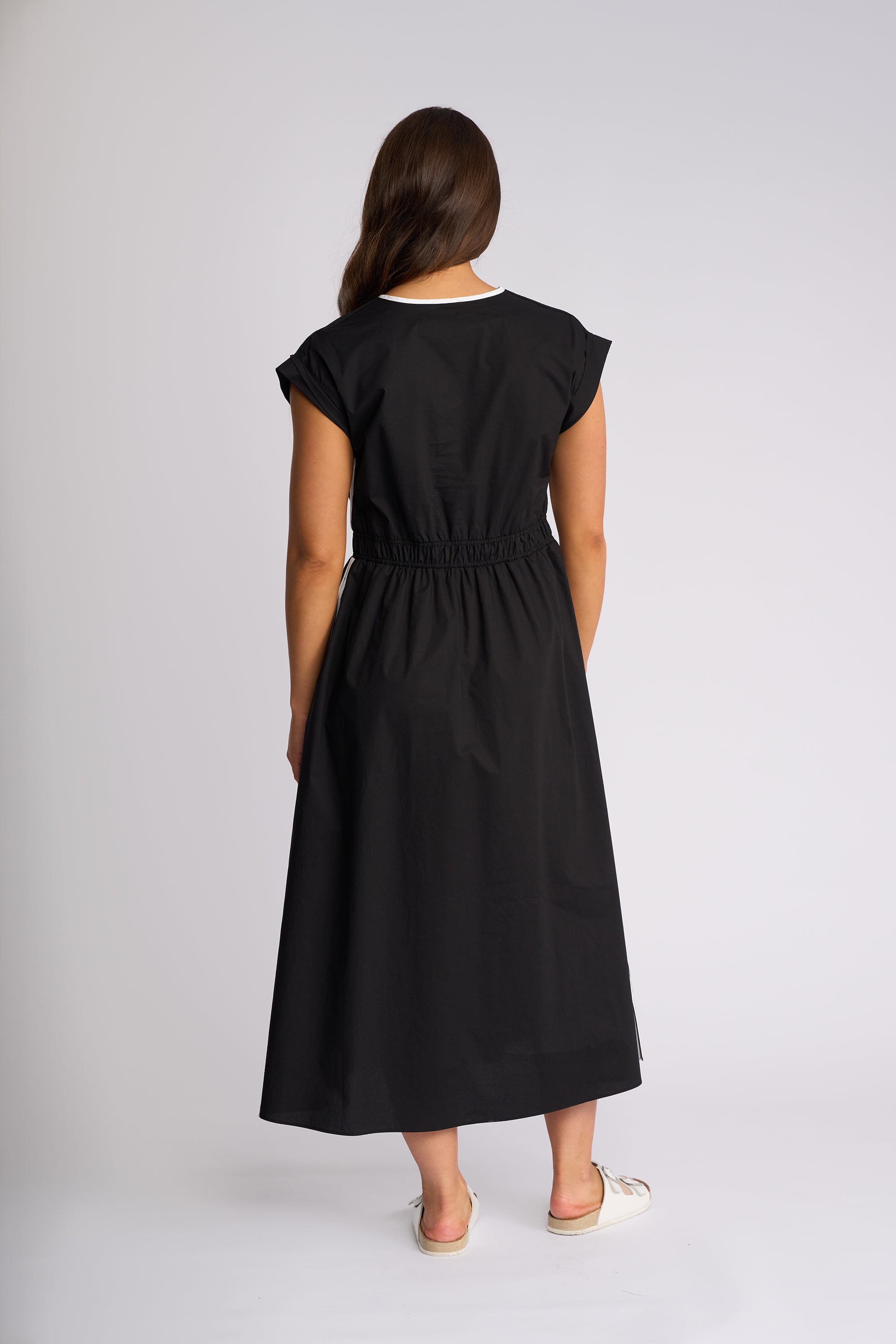 A woman viewed from the back wearing a black midi dress with cap sleeves, a gathered waist, and a white contrast collar, standing against a light gray studio background.