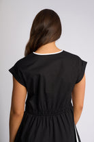 A woman viewed from the back wearing a black dress with a white contrast trim at the crew neck, set against a plain light gray studio background.