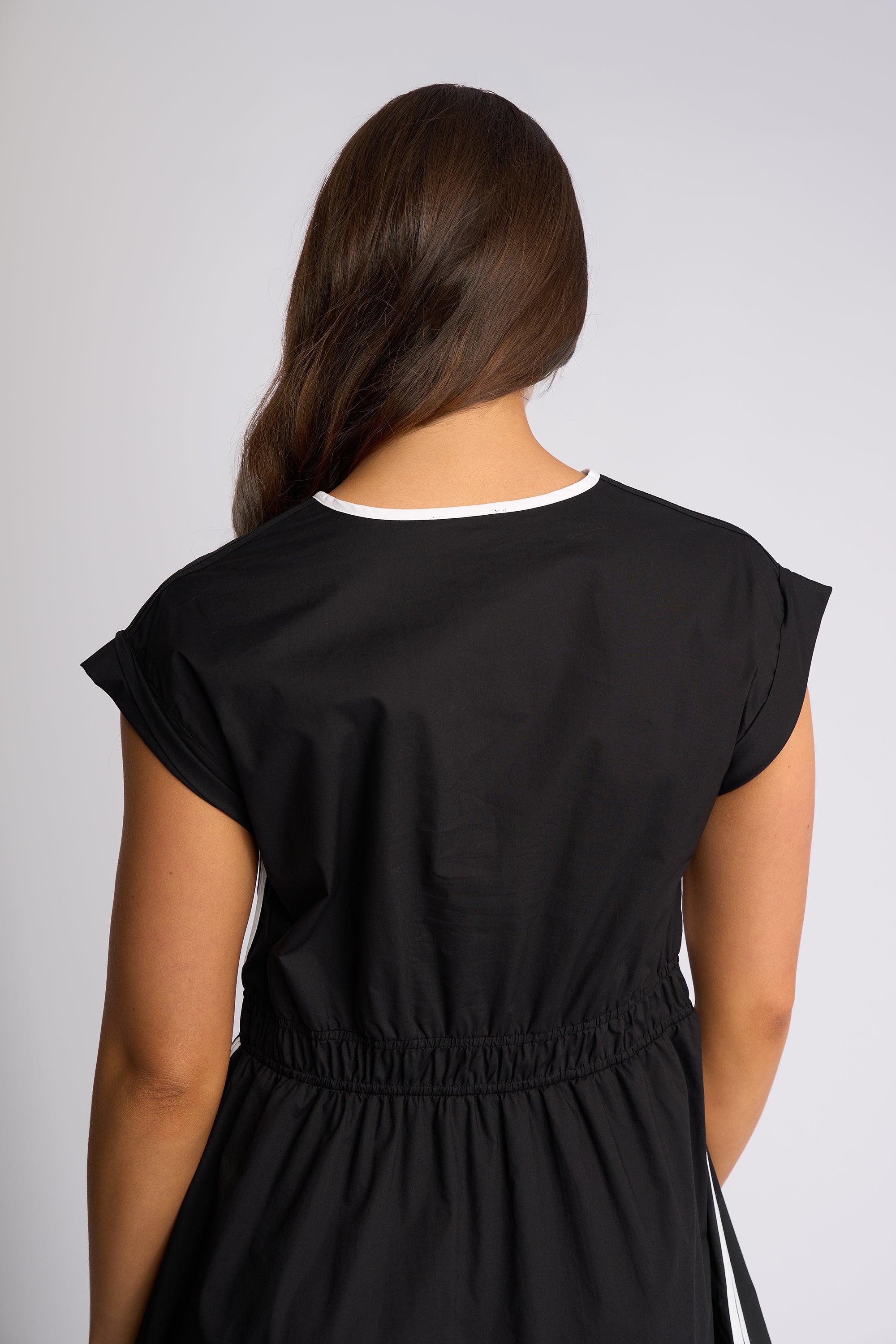 A woman viewed from the back wearing a black dress with a white contrast trim at the crew neck, set against a plain light gray studio background.