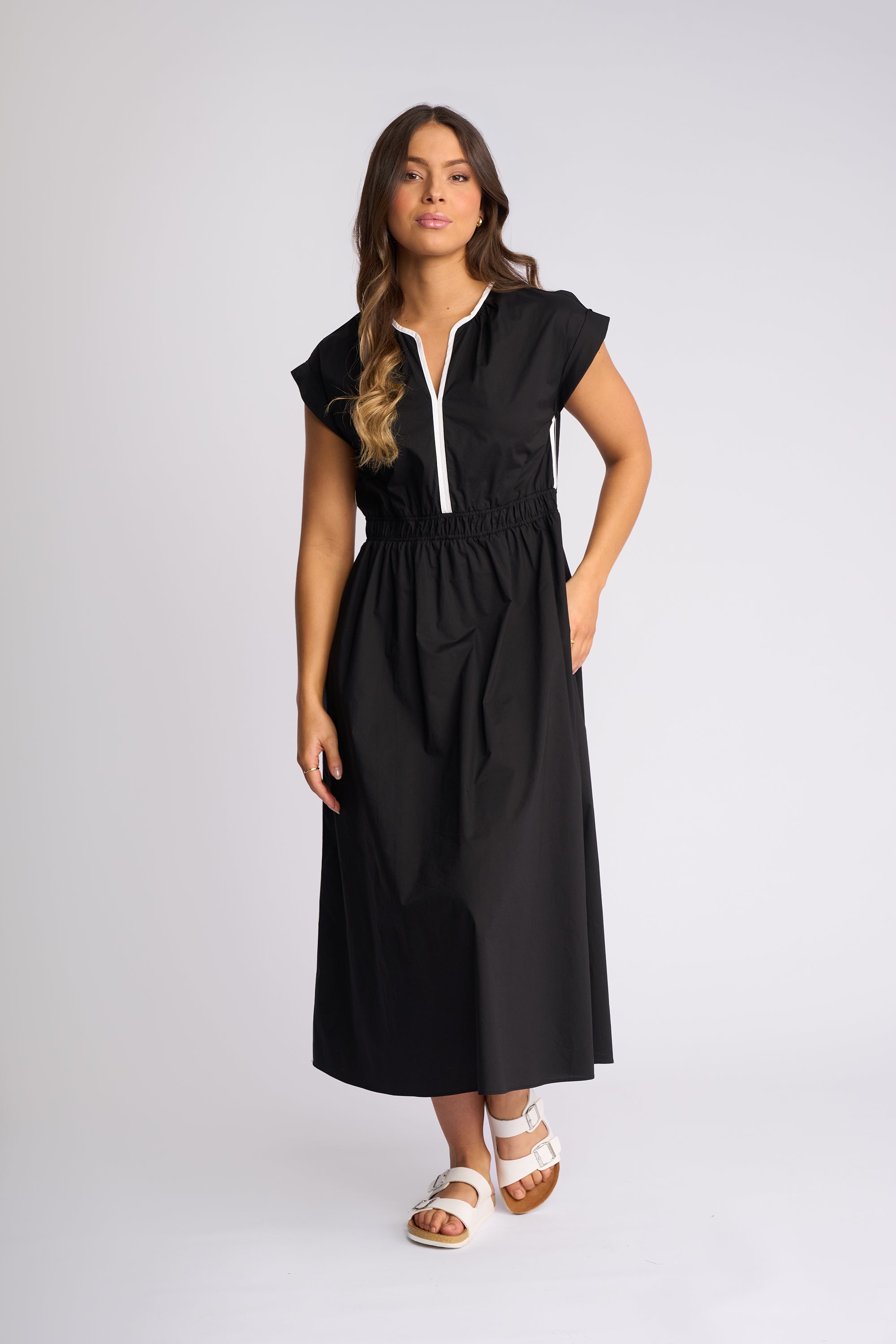 A woman modeling a black midi dress with a white zipper detail at the neckline, short cap sleeves, and side pockets, paired with white slide sandals, against a light gray studio background.