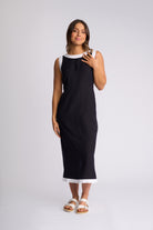 A woman modeling a sleeveless black midi dress with white contrast trim at the neckline and hem, paired with white slide sandals, against a light gray studio background.