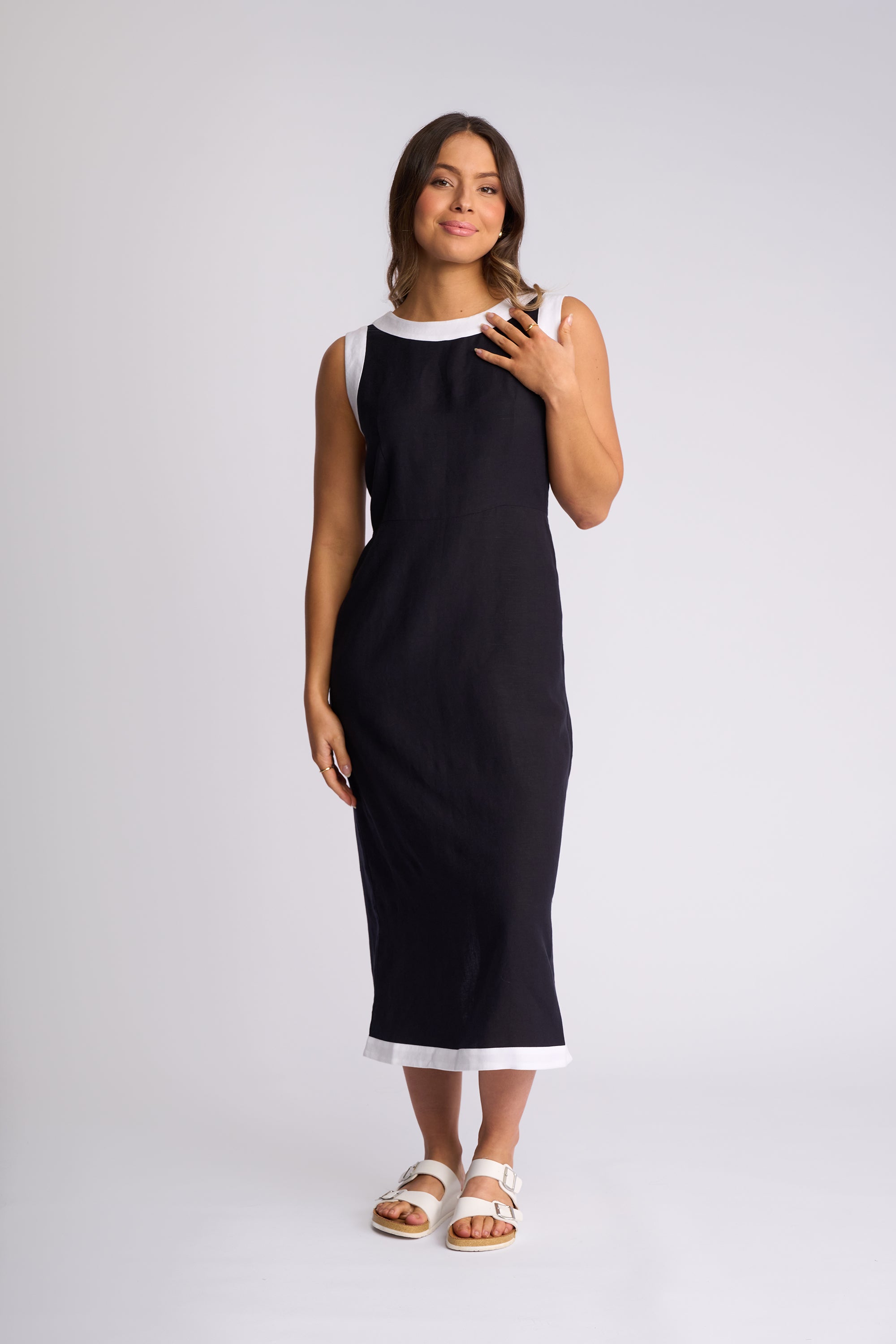 A woman modeling a sleeveless black midi dress with white contrast trim at the neckline and hem, paired with white slide sandals, against a light gray studio background.