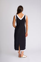 A woman viewed from the back wearing a sleeveless black midi dress with white contrast trim at the V-neckline and hem, paired with white slide sandals, against a light gray studio background.