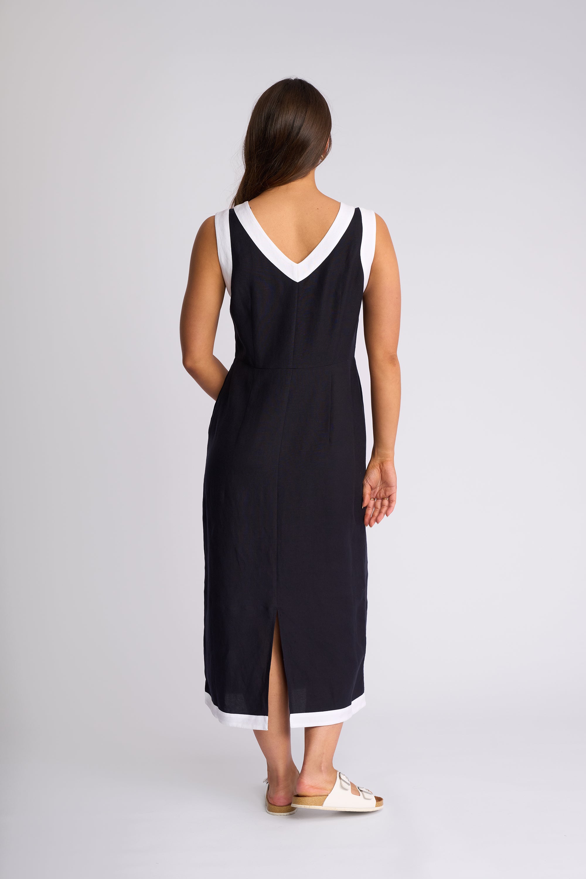 A woman viewed from the back wearing a sleeveless black midi dress with white contrast trim at the V-neckline and hem, paired with white slide sandals, against a light gray studio background.