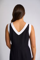 A woman viewed from the back wearing a sleeveless black dress with a contrasting white V-neck trim, set against a plain light gray studio background.