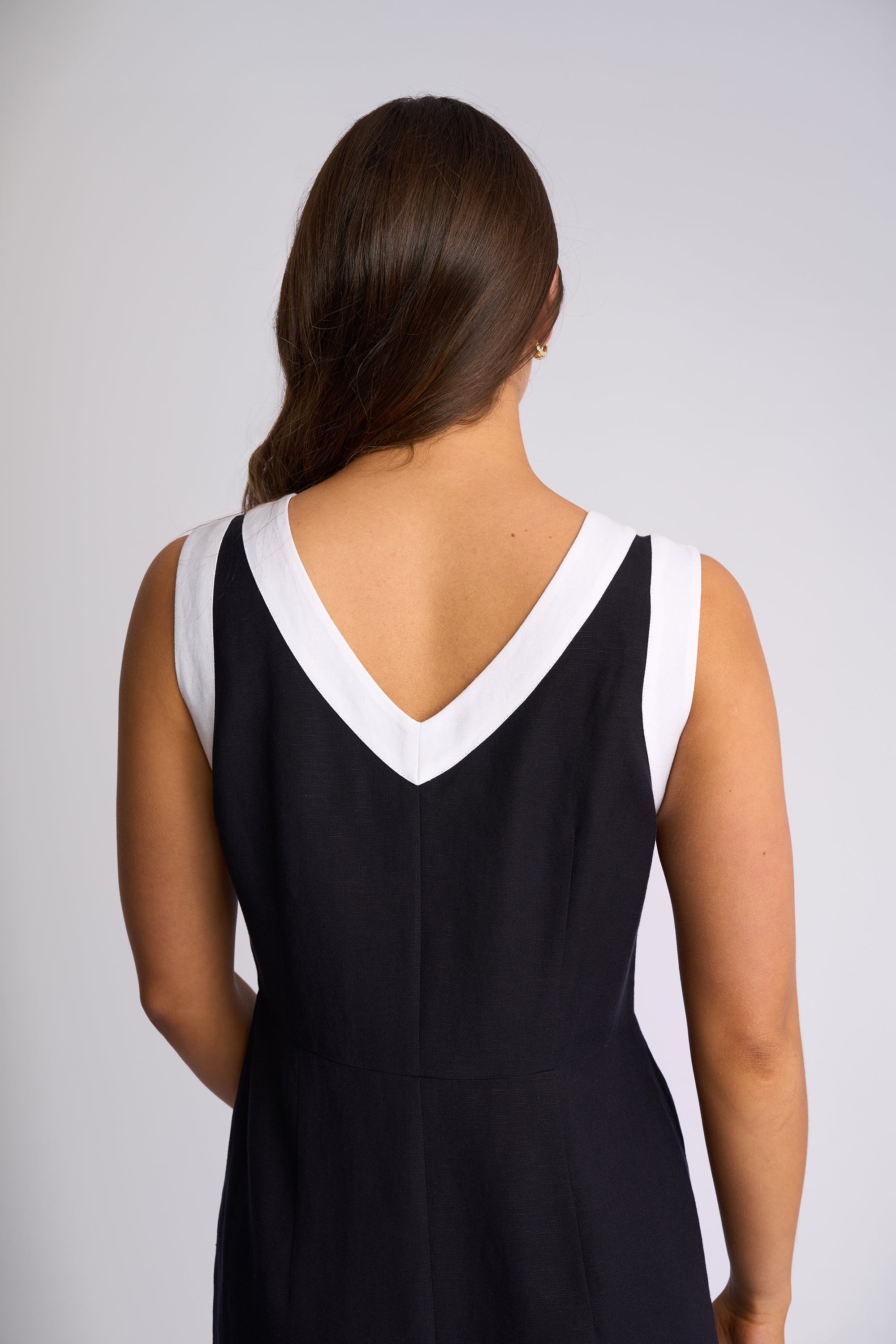 A woman viewed from the back wearing a sleeveless black dress with a contrasting white V-neck trim, set against a plain light gray studio background.