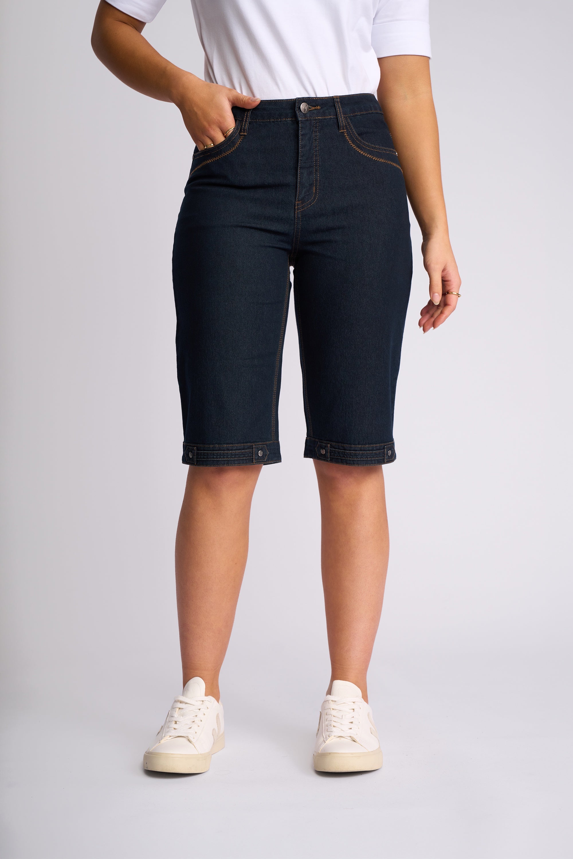 A person wearing dark navy denim biker shorts with rolled cuffs, paired with a white t-shirt and off-white sneakers, shown from the waist down against a light gray studio background.