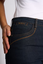 Close-up of a person wearing dark wash denim jeans with visible contrast stitching and a waistband patch reading PASSALLI. The person is also wearing a white top, and a hand with a gold ring rests near the pocket.