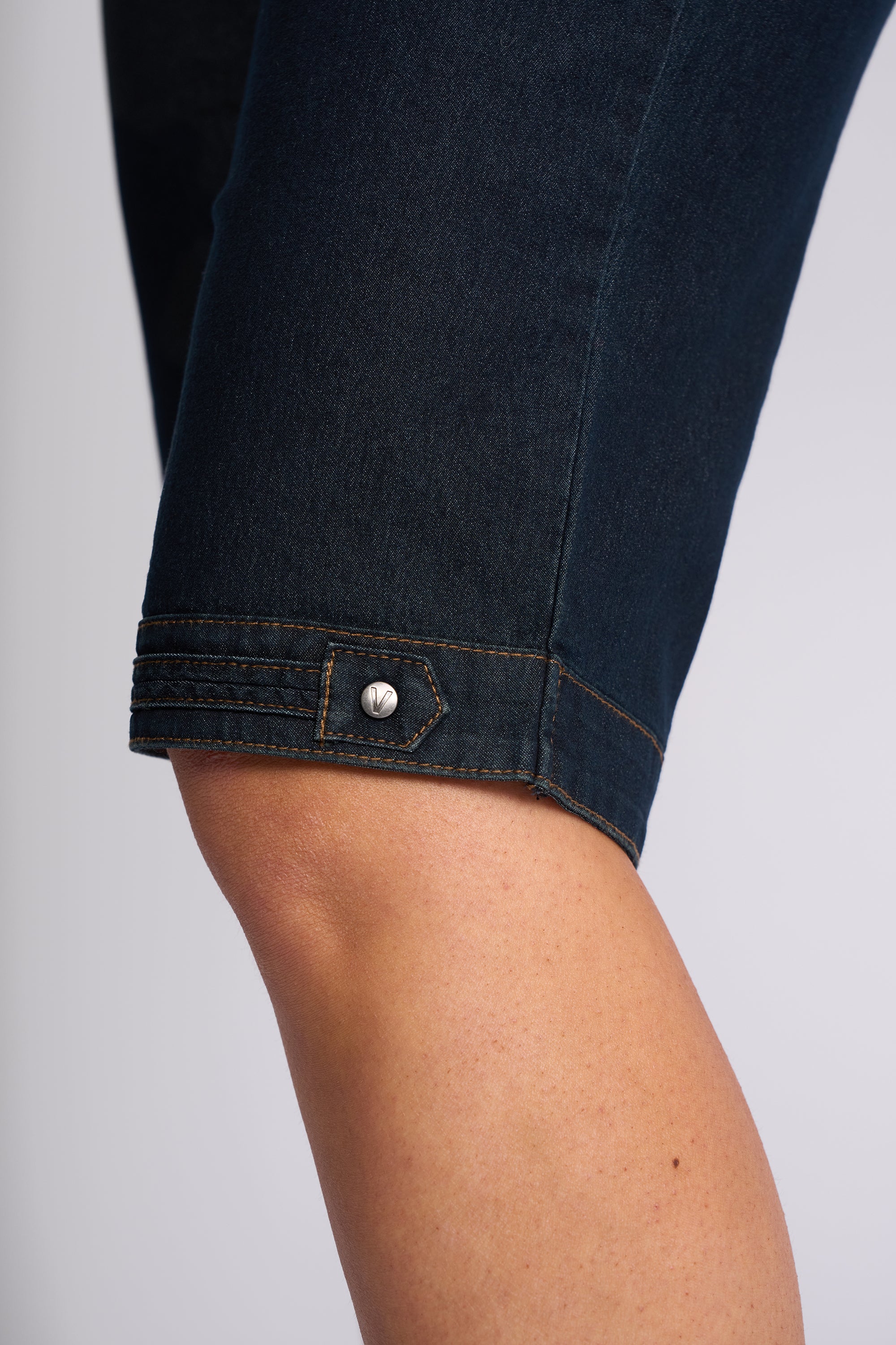 Close-up of a person's leg wearing dark indigo denim capri pants, showing the rolled cuff with a silver button detail.