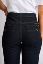 Close-up back view of a person wearing dark indigo denim jeans with contrast tan stitching and a white t-shirt, showing the back pockets and waistband.