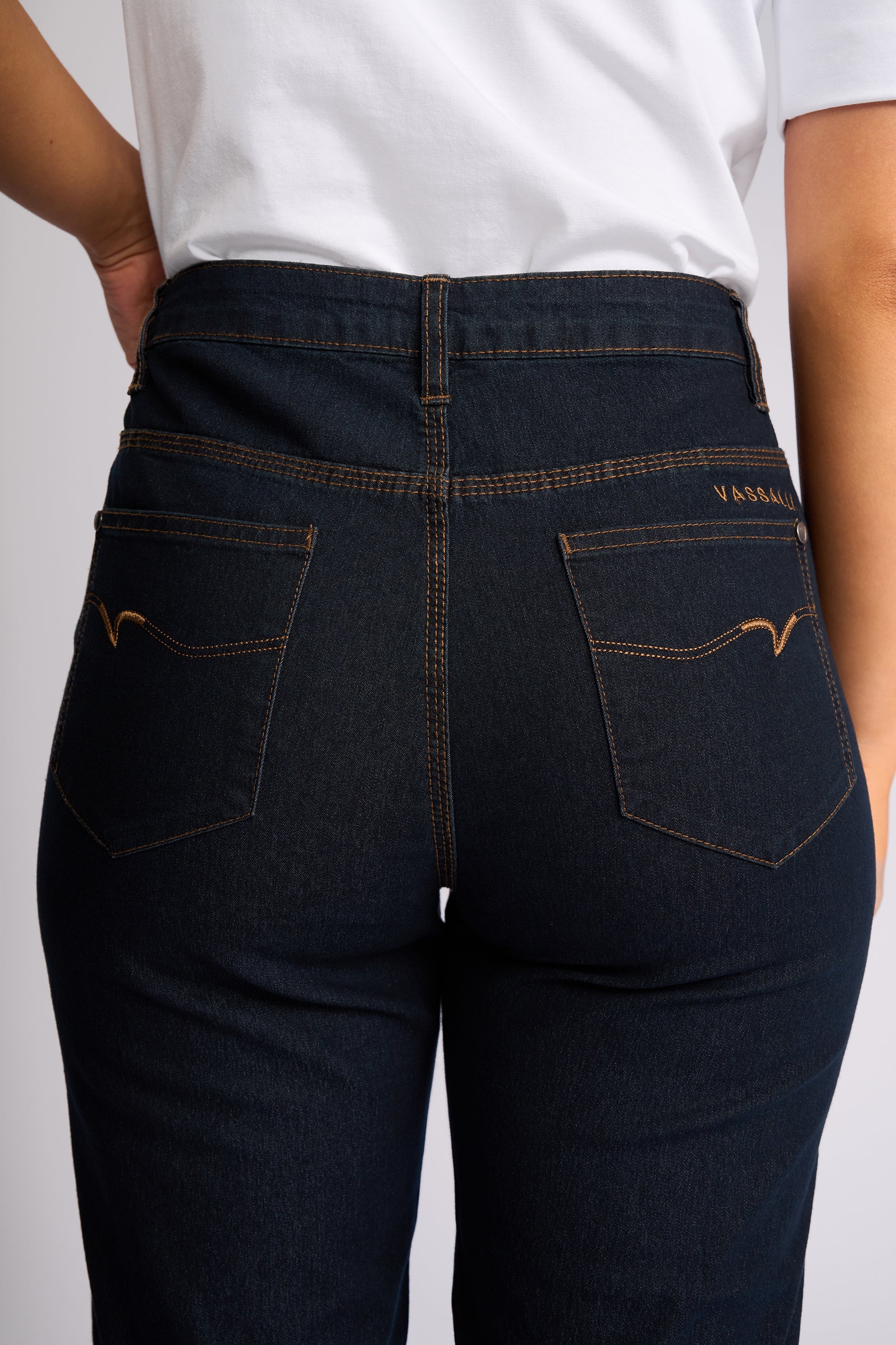 Close-up back view of a person wearing dark indigo denim jeans with contrast tan stitching and a white t-shirt, showing the back pockets and waistband.
