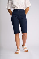 A model from the waist down wearing navy blue cropped shorts, a white short-sleeve shirt, and white slide sandals, standing against a light gray studio background.