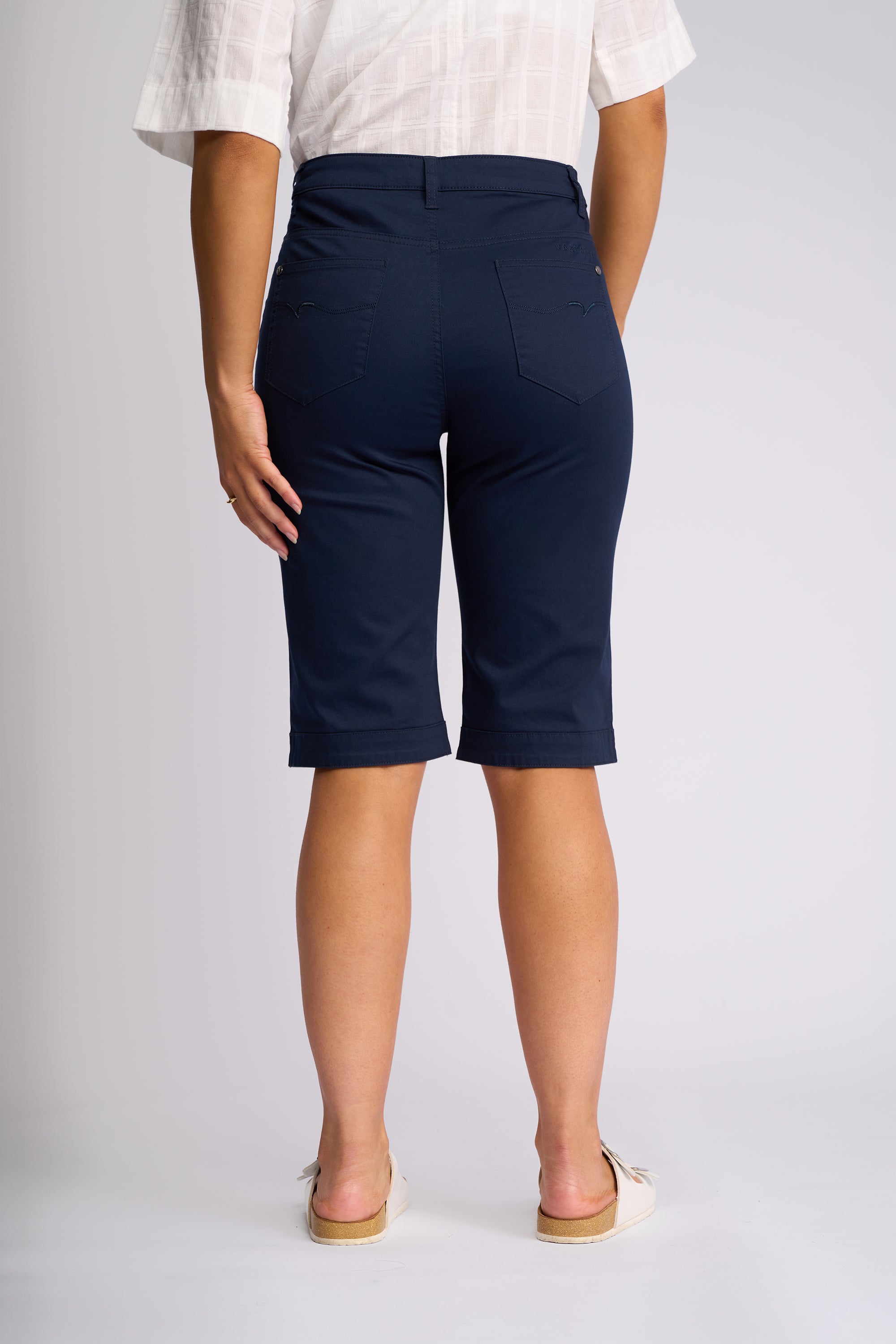 A person shown from the back wearing navy blue knee-length shorts, a white short-sleeve top, and white slide sandals on a light gray studio background.