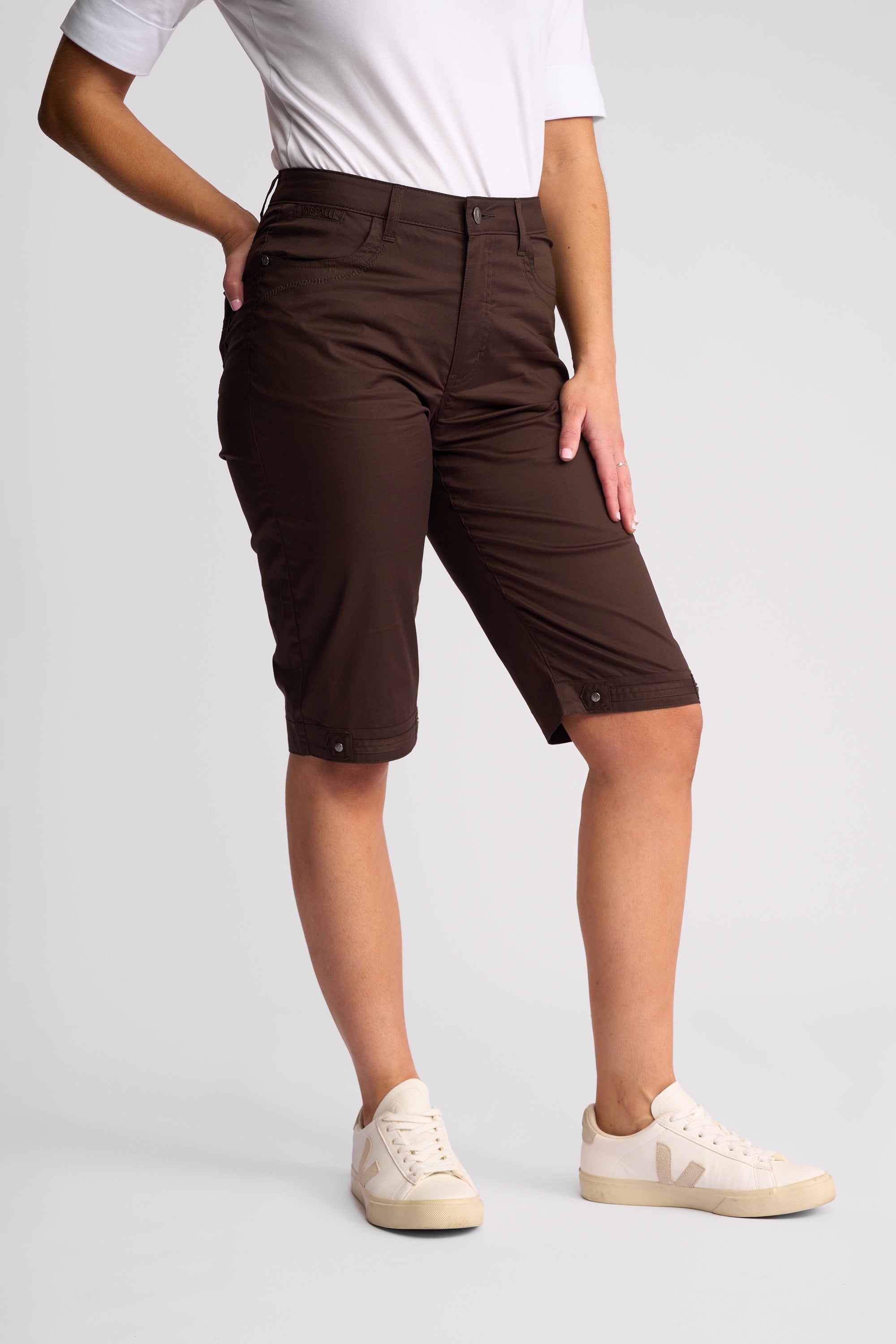 A model wearing dark brown cargo shorts, a white t-shirt, and cream-colored sneakers, standing against a light gray studio background.