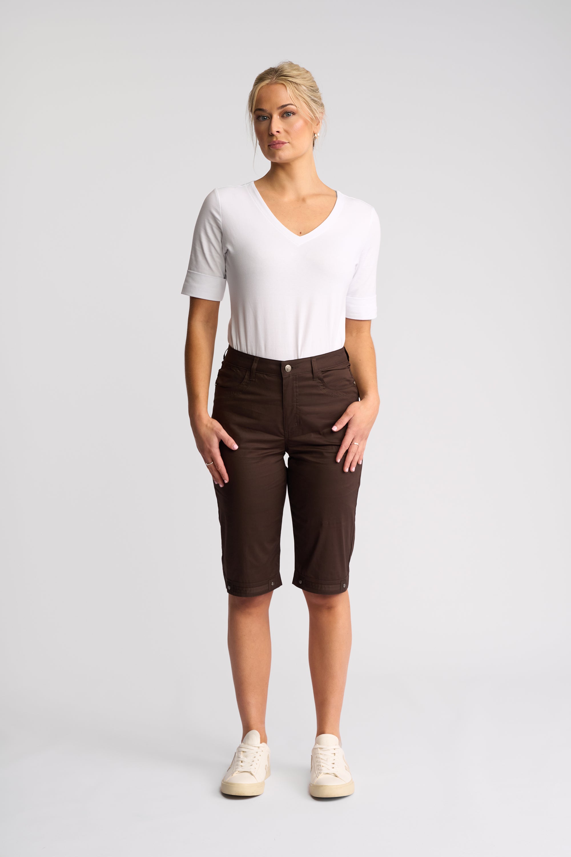 A woman modeling a white short-sleeve V-neck top and dark brown cropped pants with cream sneakers, standing against a light gray studio background.
