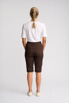 A woman viewed from behind wearing a white short-sleeve t-shirt, dark brown cropped shorts, and off-white sneakers, standing against a light gray studio background.